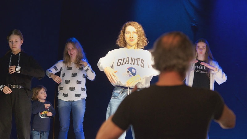Probenszene auf einer Theaterbühne mit vier jungen Frauen und einem Kind, Regisseur im Vordergrund von hinten zu sehen