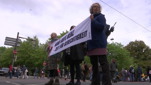 Drei Protestierende der Letzten Generation stehen auf einer Kreuzung und halten ein weißes Banner in den Händen.