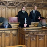 Drei Angeklagte (unkenntlich) sitzen im Gerichtssaal. Hinter ihnen stehen zwei Verteidiger.