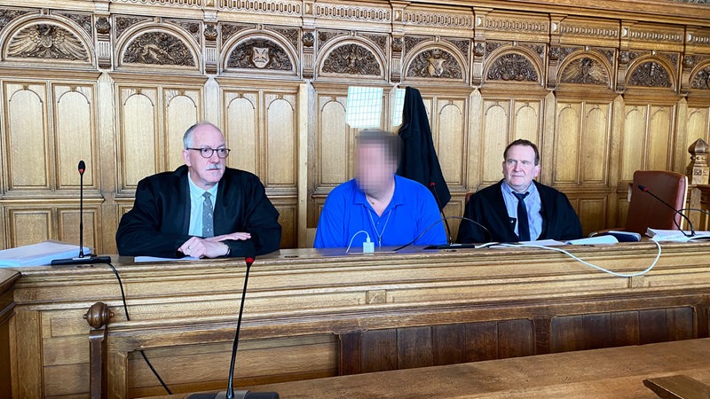 Drei Männer sitzen an einem Tisch. Das Gesicht des Mannes in der Mitte ist verpixelt. Die Männer links und rechts tragen schwarze Roben.