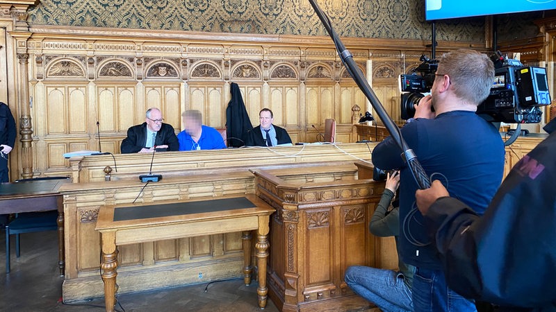 Drei Männer sitzen an einem Tisch. Das Gesicht des Mannes in der Mitte ist verpixelt. Die Männer links und rechts tragen schwarze Roben. Sie werden von einem Kameramann gefilmt.
