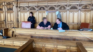 Zwei Männer sitzen an einem Tisch. Das Gesicht des linken Mannes ist verpixelt. Im Hintergrund ist eine Frau in Justizuniform zu sehen.