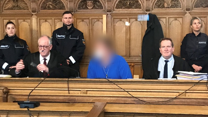 Ein Mann im blauen T-Shirt sitzt auf der Anklagebank. Sein Gesicht ist verpixelt. Links und rechts neben ihm sitzen seine beiden Anwälte. Hinter ihm stehen drei Justizbeamte.