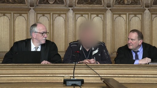 Drei Personen in einem Gericht, das Gesicht der mittigen Person wurde unkenntlich gemacht.