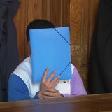 Ein Angeklagter im weißen Pullover sitzt in einem Gerichtssaal, eine blaue Mappe vor dem Gesicht