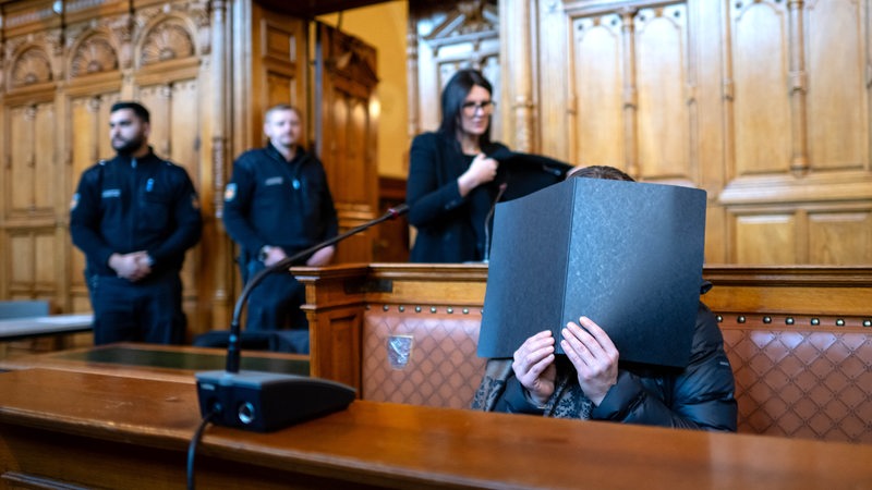 Eine Angeklagte sitzt vor Prozessbeginn im Gerichtssaal, vor ihrem Gesicht hält sie eine Akte.