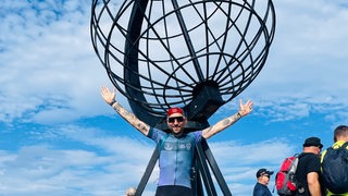 Extrem-Radsportler Florian Schigelski steht strahlend mit ausgebreiteten vor der Globus-Skulptur bei seiner Ankunft am Nordkap.