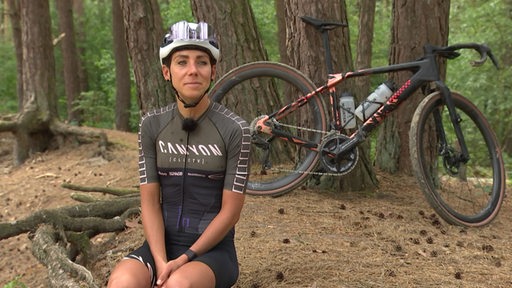 Radprofi Carolin Schiff sitzt in einem Wald. Im Hintergrund ist ihr Rennrad zu sehen, welches an einen Baum lehnt.