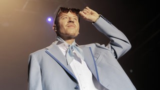 Ein Mann im Sakko steht auf einer Bühne und hebt seine Sonnenbrille an.