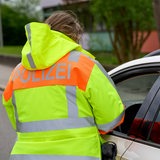 Eine Frau, die eine gelb-orange Jacke mit der Aufschrift "Polizei" auf dem Rücken trägt, steht neben einem Auto. Das Fenster am Fahrerplatz ist geöffnet.