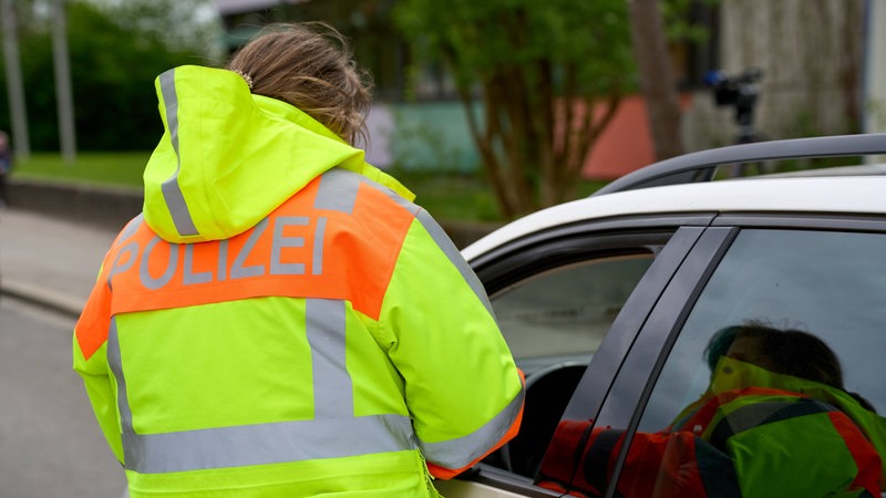 Eine Frau, die eine gelb-orange Jacke mit der Aufschrift "Polizei" auf dem Rücken trägt, steht neben einem Auto. Das Fenster am Fahrerplatz ist geöffnet.