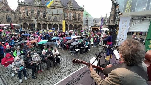 Blick von einer Bühne auf den Rathausplatz, wo viele Menschen mit Regenschirmen die Welterben (Roland und Rathaus) feiern. 