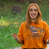 Eine junge Frau mit rötlichen Haaren und einem orangefarbenen T-Shirt ist im Vordergrund zu sehen, im Hintergrund ist eine Ratte zu sehen.