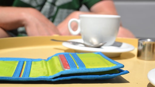 Ein grün-blaues Portemonnaie liegt geöffnet auf einem Tisch. Dahinter sitzt ein Mensch vor einer weißen Kaffeetasse. Nur die Hände sind unscharf sichtbar.