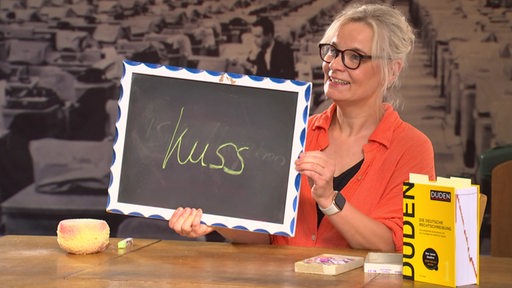 Eine Frau sitzt an einem Tisch und hält eine kleine Tafel hoch. Auf der Tafel steht das Wort "Kuss" geschrieben. 