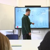 Zu sehen ist ein junger angehender Lehrer, welcher vor einer Klasse unterrichtet.
