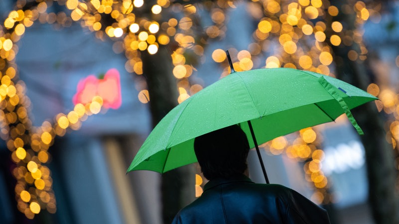 Ein Fußgänger läuft mit einem grünen Regenschirm durch eine weihnachtlich beleuchtete Straße.