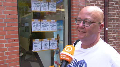 Ein Mann mit Glatze und Brille steht vor einem Reisebüro und wird interviewt.