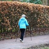 Eine Frau läuft auf einem Weg neben einer Hecke mit braunen Bättern. Sie trägt eine hellblaue Jacke, eine schwarze Hose und läuft mit zwei Nordic-Walking-Stöcken-