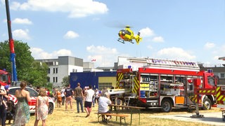 Auf einem Platz sind Menschen und ein Feuerwehrfahrzeug zu sehen. Am Himmel fliegt ein Rettungshubschrauber.