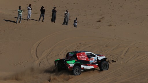 Ein paar Zuschauer stehen in einem Wüstenstück in Saudi Arabien, während ein Auto der Rallye Dakar passiert.
