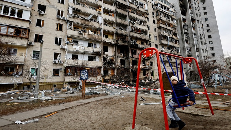 Ein Kind sitzt auf einer Schaukel vor einem zerstörten Wohnhaus in Kiew