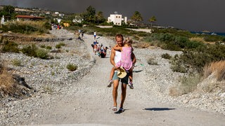 Rhodos: ein Mann trägt ein Kind auf dem Arm, als sie ein Waldbrandgebiet verlassen müssen.