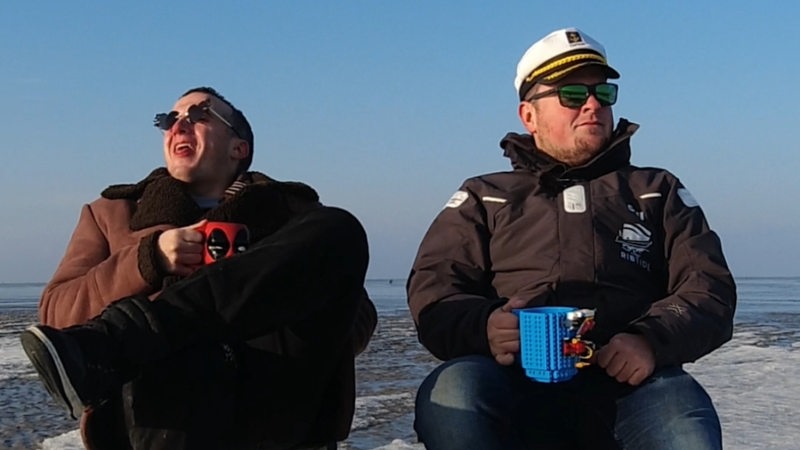 Das Duo "Ribtide" sitzen mit Kaffeetassen auf Klappstühlen vor Bremerhavener Kulisse