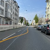 Eine gelbe Linie führt in Schlangenlinien über eine Straße. Links sind Häuser zu sehen, rechts stehen Autos.