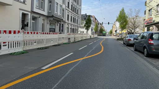 Eine gelbe Linie führt in Schlangenlinien über eine Straße. Links sind Häuser zu sehen, rechts stehen Autos.