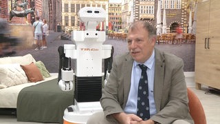 Michael Beetz vom Institut für künstliche Intelligenz im Robotic Apartment, im Hintergrund ein Roboter namens Tiago im Robotic Apartment.
