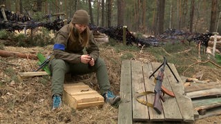 Eine Soldatin sitzt im Wald, neben ihr eine Waffe.