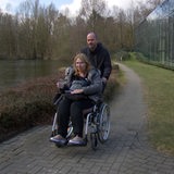 Ein Mann schiebt eine Frau im Rollstuhl auf einem Weg neben einem Gewässer.
