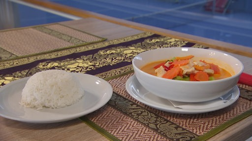 Ein rotes Curry mit Huhn und Gemüse steht neben einem Teller Reis, im Hintergrund ein Tennisplatz