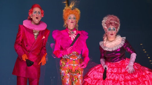 Drei Schauspieler in rot und pinkfarbenen Kostümen auf einer Bühne.