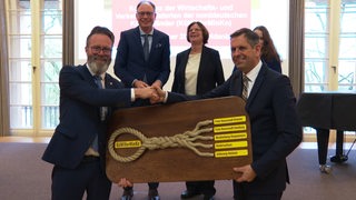 Die Wirtschafts- und Verkehrsminister Norddeutschlands stehen zusammen, zwei schütteln sich die Hande und halten ein Holzschild