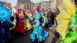 Frau im blau-grünen Sambakleid mit Blumen-Hut tanzt vor ihrer Gruppe neben dem Bremer Dom.