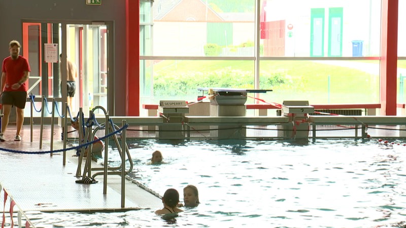 Im Innen-Schwimmbecken sind mehrere Menschen im Wasser. Am Beckenrand läuft ein Bademeister.