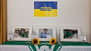 Geschmückter Tisch mit  Werder-Flagge, zwei Sonnenblumen und drei Bilderrahmen mit Fotos der drei ukrainischen Werder-Schachspieler, die am Saisonauftakt nicht teilnehmen konnten.