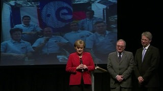 Angela Merkel steht vor einer Videowand, die sechs Astronauten im All zeigt
