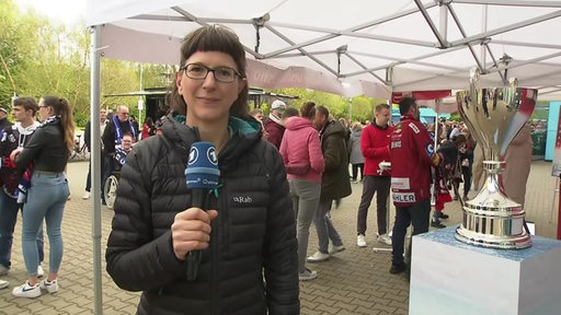 Die Sportblitz-Reporterin Maike Albrecht moderiert vor der Eisarena in Bremerhaven. Neben ihr steht ein Pokal auf einem Sockel.
