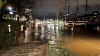 Treppenstufen, eine Mauer und Wasser, das die Stufen überschwemmt. Im Hintergrund Schiffe.
