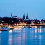 Die Lichter der Uferpromenade Schlachte spiegeln sich in der Dämmerung in der Weser, während im Hintergrund die Altstadt und der Bremer Dom St. Petri zu erkennen sind.