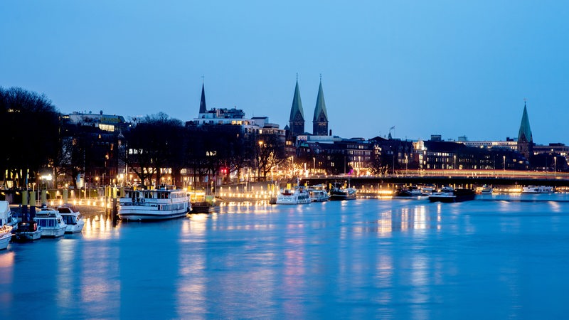 Die Lichter der Uferpromenade Schlachte spiegeln sich in der Dämmerung in der Weser, während im Hintergrund die Altstadt und der Bremer Dom St. Petri zu erkennen sind.