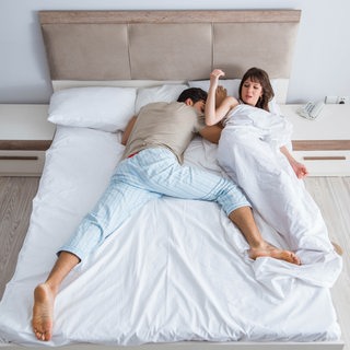 Eine Frau liegt sichtlich genervt neben einem schlafenden Mann in einem Doppelbett.