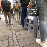 Menschen stehen in einer Schlange - eine Frau trägt einen mobilen Sitz mit sich