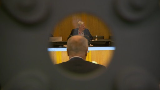 Der Blick durch ein rundes Schlüsselloch, man sieht zwei Männer in einem Saal.