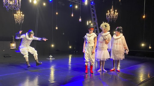 Ein Clown sitzt auf einer Schaukel auf einer Bühne, daneben stehen drei weitere Clowns.