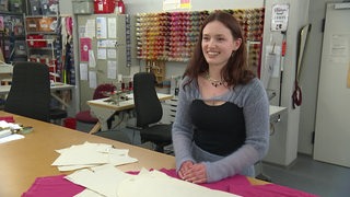 Eine junge Frau sitzt in einer Werkstatt am Tisch, auf dem Schablonen und Stoffe liegen