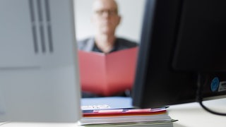 Ein Mann sitzt hinter Computerbildschirmen und liest in einem Ordner.